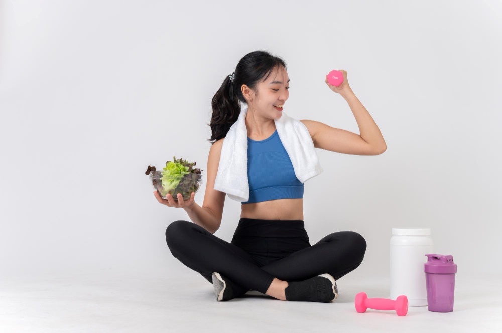 หญิงที่กำลังนั่งขัดสมาธิสวมเสื้อกีฬา ในมือซ้ายถือชามสลัดและในมือขวาถือลูกเหล็กสีชมพูขนาดเล็ก 